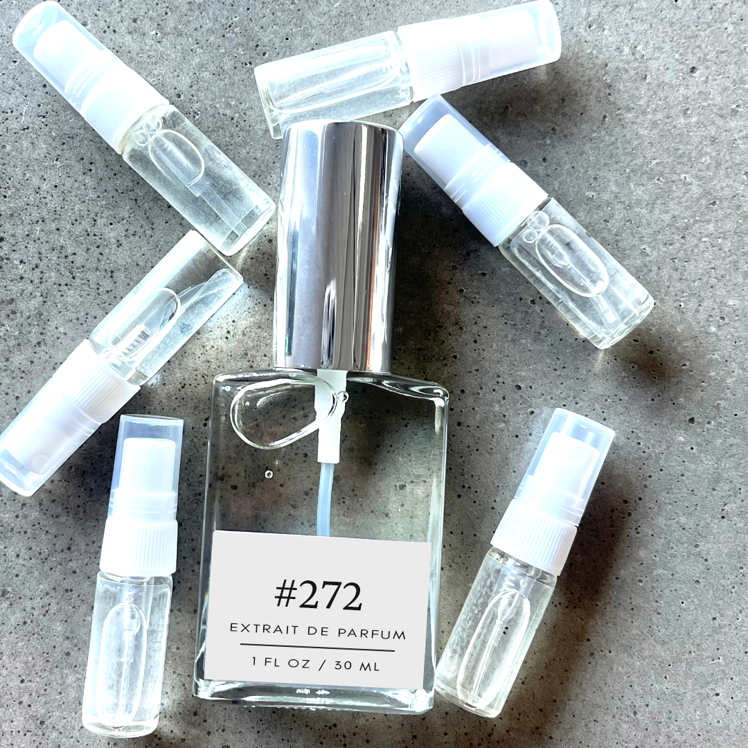Clear bottle labeled with #272 Extrait De Parfum with silver cap, accompanied by 6 elegantly arranged sample bottles, resting on a marble surface.