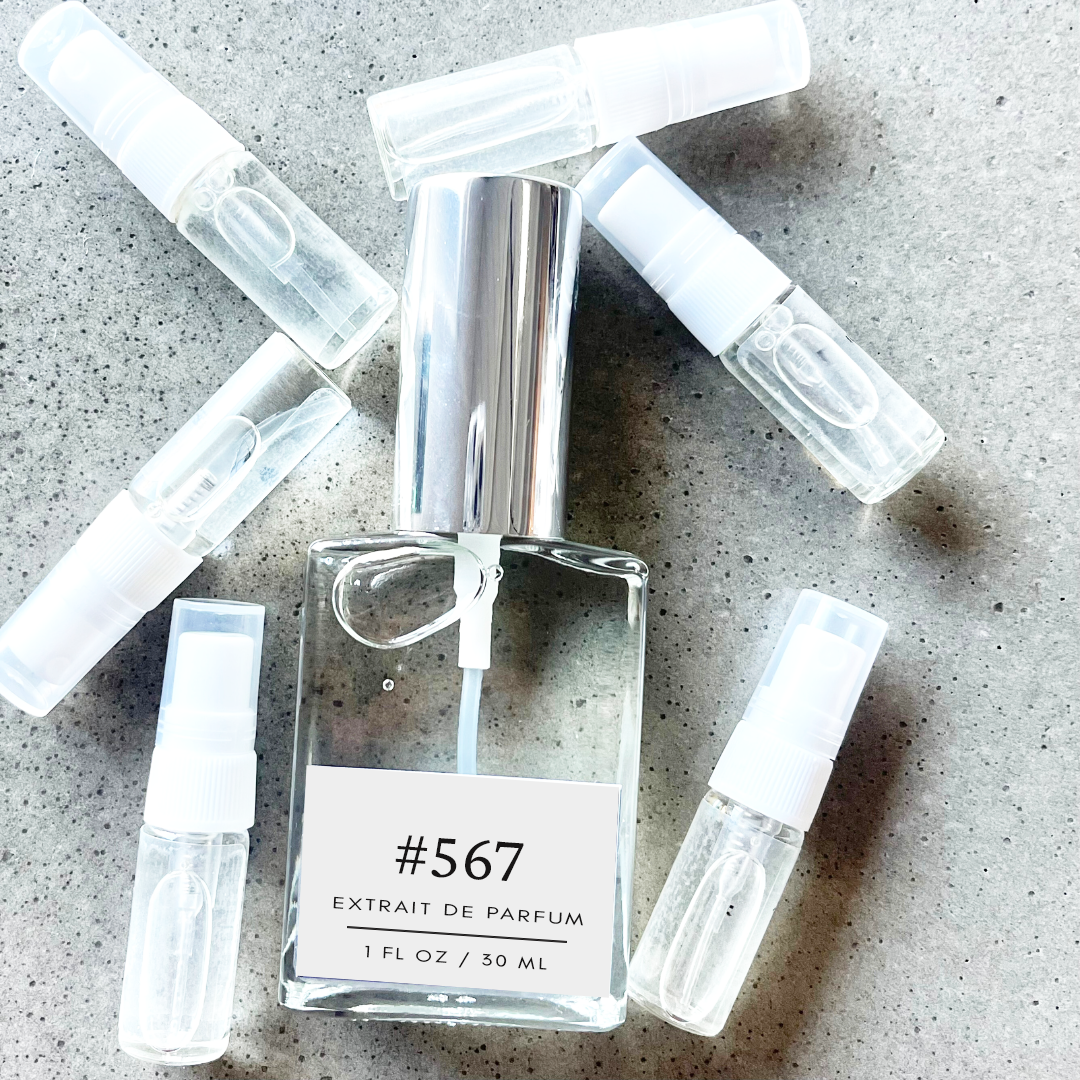 Clear bottle labeled with #567 Extrait De Parfum with silver cap, accompanied by 6 elegantly arranged sample bottles, resting on a marble surface.