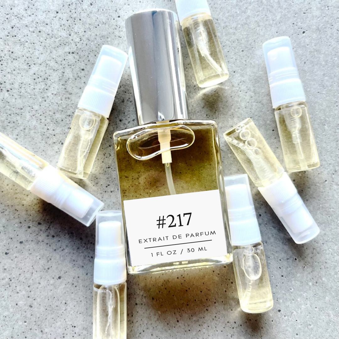 Clear bottle labeled with #217 Extrait De Parfum with silver cap, accompanied by 7 elegantly arranged sample bottles, resting on a marble surface