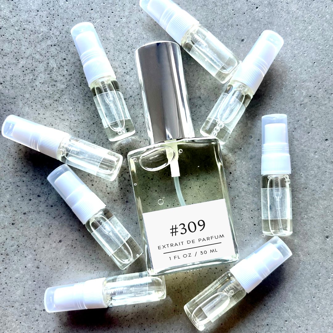 Clear bottle labeled with #309 Extrait De Parfum with silver cap, accompanied by 8 elegantly arranged sample bottles, resting on a marble surface.